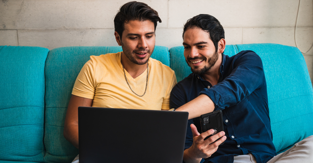 Two Hispanic men sitting on a couch engaging with digital content on a laptop and smartphone, representing modern Hispanic consumer behavior and online connectivity.