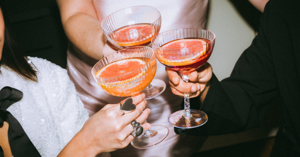 Three stylish Gen Z individuals clink coupe glasses filled with vibrant cocktails garnished with grapefruit slices, capturing a trendy, social moment that reflects the generation’s preference for visually appealing and shareable drink experiences.