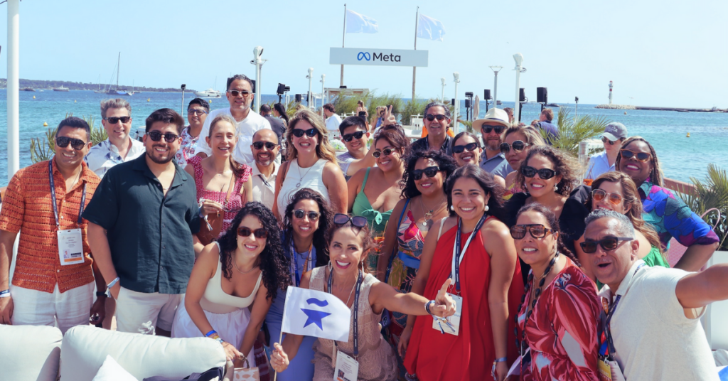 A vibrant group of diverse creatives and marketing professionals pose together on a sunny boardwalk at the Cannes Lions Festival, with the Meta beach lounge and the turquoise sea in the background.