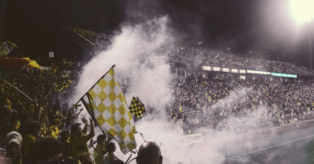 Packed stadium with diverse fans waving yellow checkered flags and celebrating amid smoke effects during a nighttime soccer match.