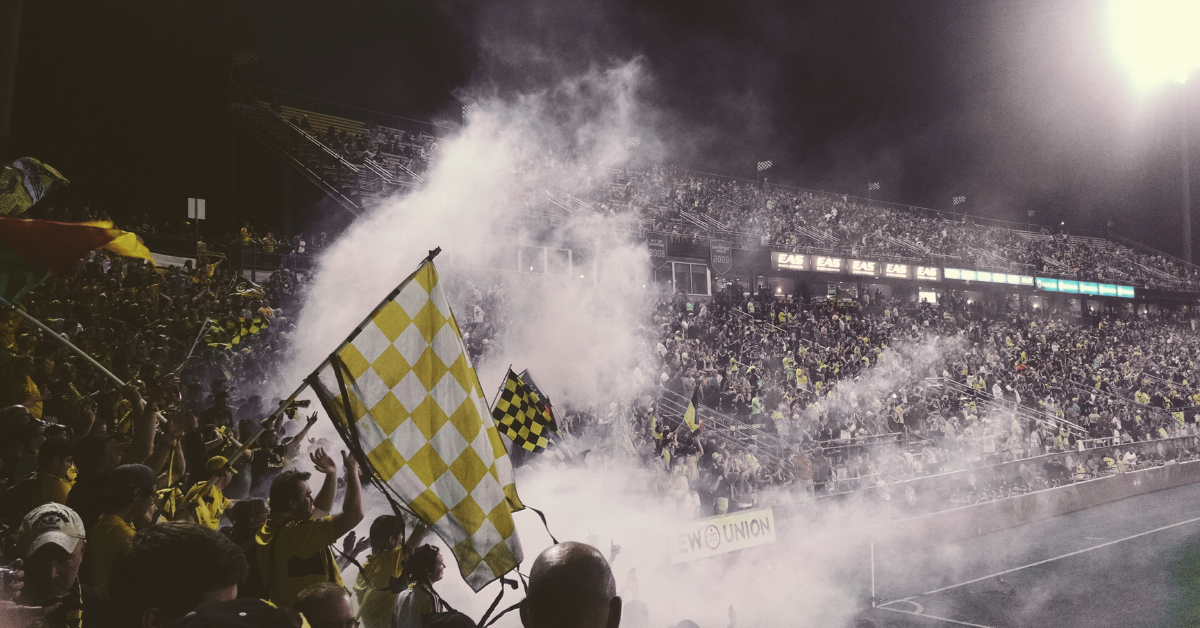 Packed stadium with diverse fans waving yellow checkered flags and celebrating amid smoke effects during a nighttime soccer match.